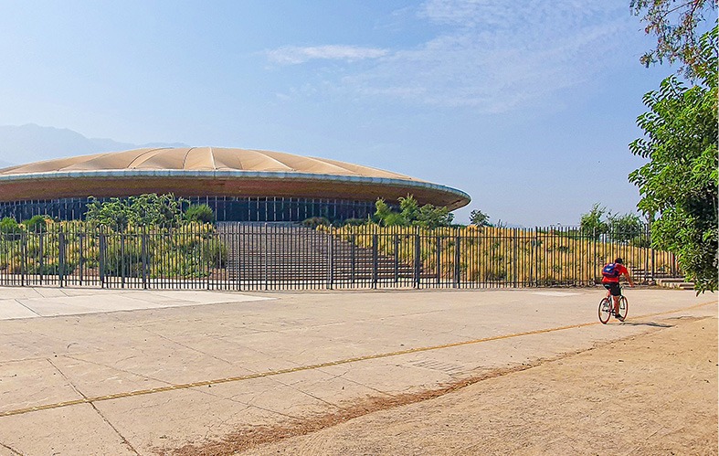 On the exterior image we can see a central shot of 
                                    the velodrome of Peñalolen from the exterior side 
                                    of the park, appreciating the lattice that surrounds 
                                    the area and a person that passes by on their bike.